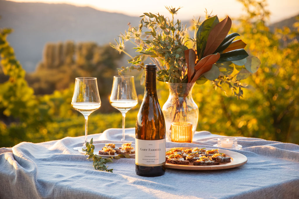 A bottle of Gary Farrell 2023 Russian River Chardonnay with two wine glasses on a simple table scape with a Fall Crostini and a sunset backdrop.