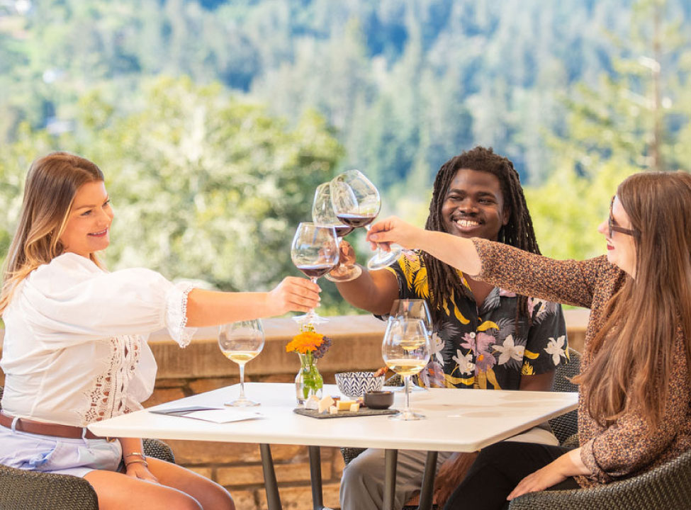 Three friends clink glasses together at Gary Farrell Winery with a view of the Russian River Valley behind them.
