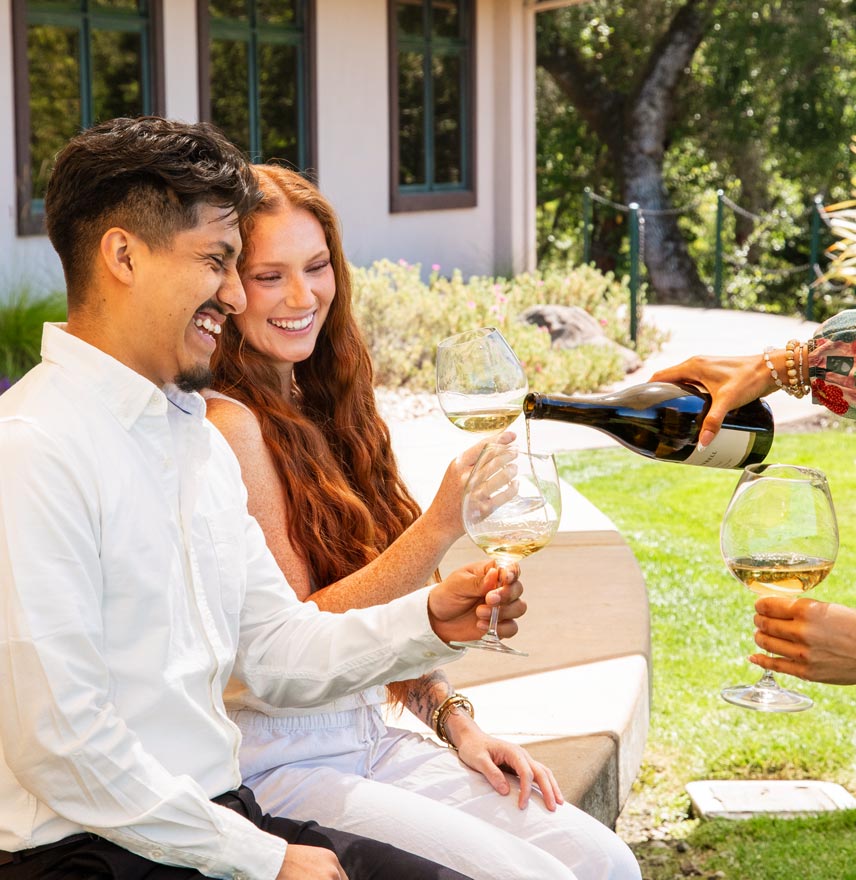 A man and woman sit on the lawn at Gary Farrell Winery in Healdsburg, CA. They hold up their wine glass for a fresh pour of Russian River Valley Chardonnay.